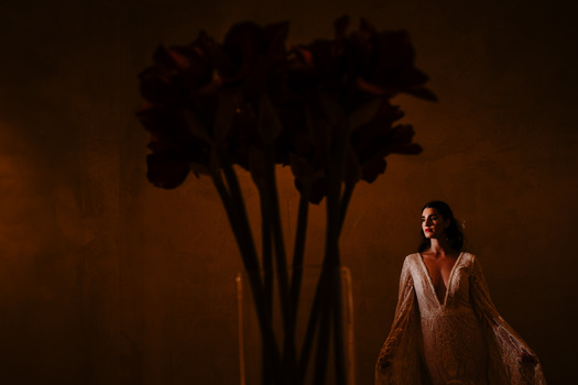 Woman in beige textured garment standing beside dark floral arrangement against warm brown backdrop.