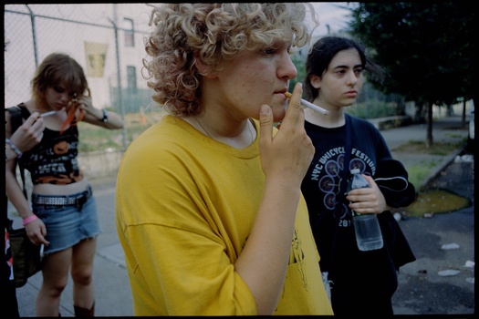 Young man in yellow shirt with curly hair standing in outdoor setting with other teenagers nearby.
