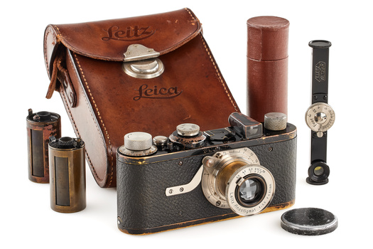 Vintage Leica rangefinder camera with leather case, light meters, film canisters, and wristwatch displayed on black surface.