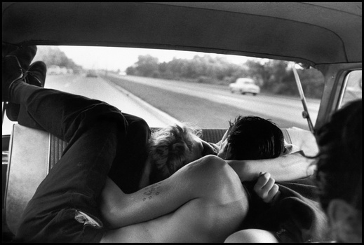 Two people in a car viewed from behind, with a highway and vehicles visible through the windshield.