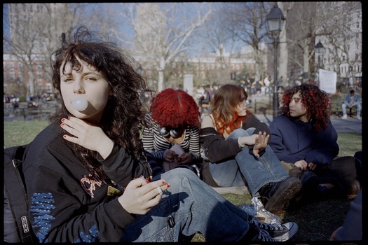Young person with dark curly hair smoking in a park with friends seated nearby.