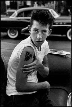 Young man in white t-shirt with shoulder tattoo sitting in front of classic car.