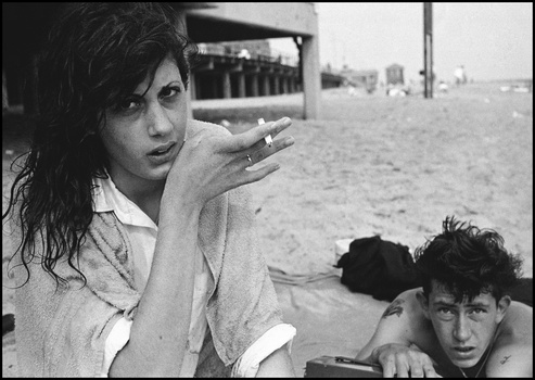 Woman smoking a cigarette on a beach, looking toward camera while seated next to a shirtless man.