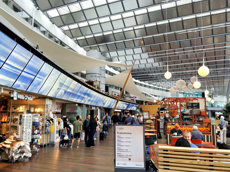 Modern airport or transit terminal interior with geometric ceiling grid, retail storefronts, and scattered pedestrians during daytime.