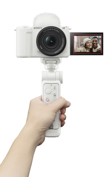 Hand holding a compact gimbal stabilizer with mirrorless camera mounted on top and flip-out LCD screen visible.