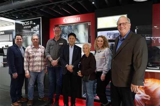 Seven people posing together in front of Canon and camera equipment displays at what appears to be a photography trade show or exhibition.
