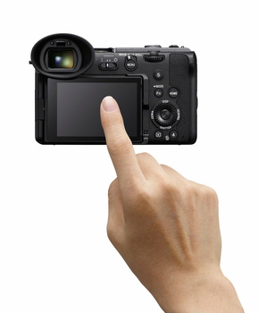 Hand pointing at the rear LCD screen of a mirrorless camera against white background.