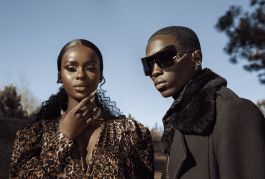 Two people pose outdoors against a clear sky, one wearing a leopard print shirt and the other in a black jacket with sunglasses.