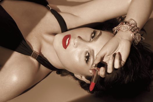 Woman with red lipstick and bold eye makeup tilting head back against neutral background.