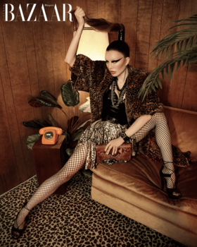 Model in sequined jacket and fishnet stockings posed on beige chair in wood-paneled interior.