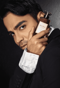 Man in dark suit holding amber-colored fragrance bottle near face against dark background.