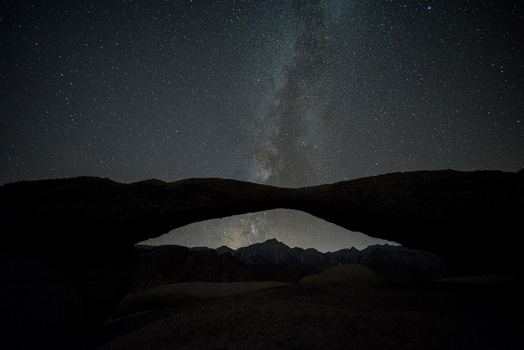 Milky Way galaxy visible through a natural rock arch at night, with silhouetted mountains and desert landscape.