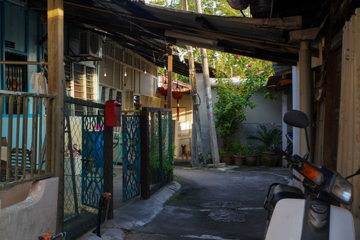 Narrow alleyway with blue metal gates, wooden structures, and dappled sunlight filtering through overhead beams.