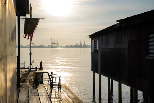 Silhouetted wooden structures on a dock with industrial cranes visible across calm water at sunrise or sunset.