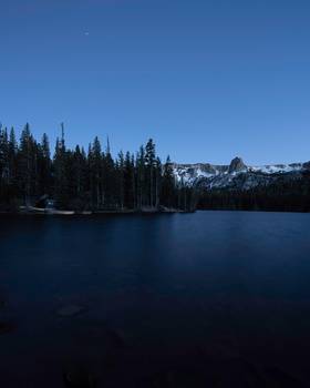 Alpine lake at dusk with coniferous forest and snow-capped peaks reflected in still water.
