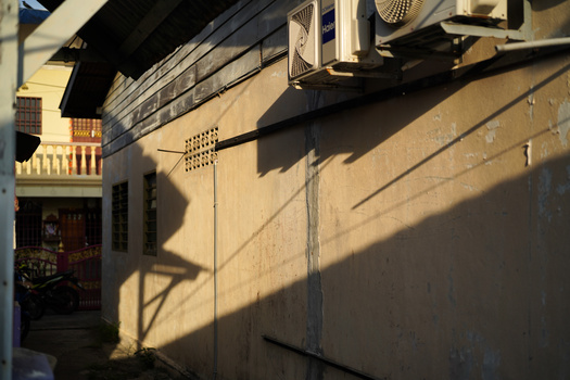 Strong shadow patterns cast by an air conditioning unit and overhead structure on a cream-colored building wall.
