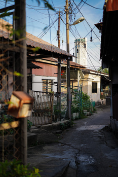Narrow alleyway with weathered buildings, power lines, and a bridge visible overhead in warm sunlight.