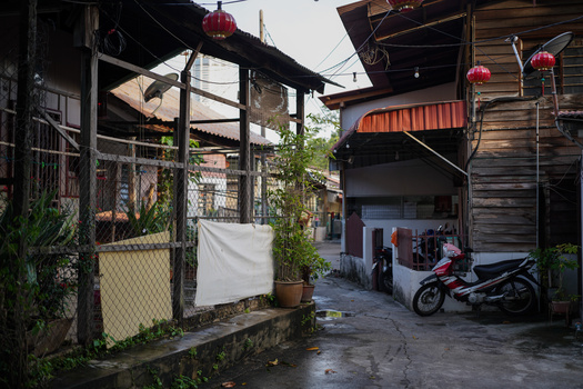 Narrow alleyway between weathered wooden structures with motorbike and laundry visible.