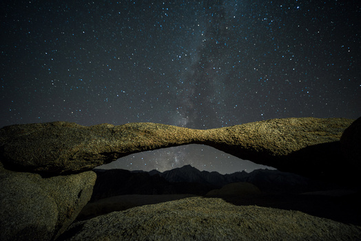 Milky Way galaxy visible through a natural stone arch at night, with mountains silhouetted in the distance.