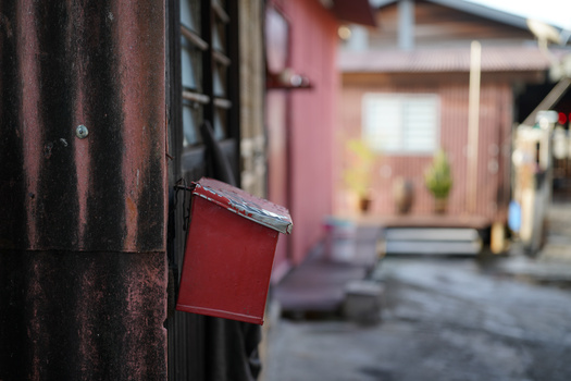 Red mailbox mounted on a dark wooden post with a blurred residential alley in the background.