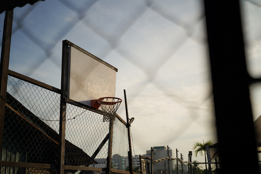 Basketball hoop and backboard on outdoor court viewed through chain-link fence and structural frame.