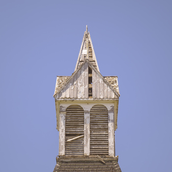Weathered wooden church steeple with louvered belfry against clear blue sky.