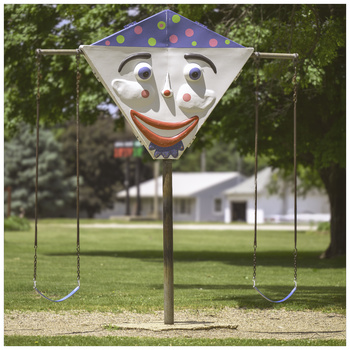 Whimsical clown face decoration mounted on wooden pole in grassy park setting.