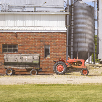 Red vintage tractor parked beside a wooden farm wagon in front of a brick agricultural building with metal grain silos.