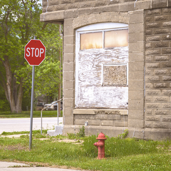 Abandoned brick building with boarded window, fire hydrant, and stop sign in overgrown lot.