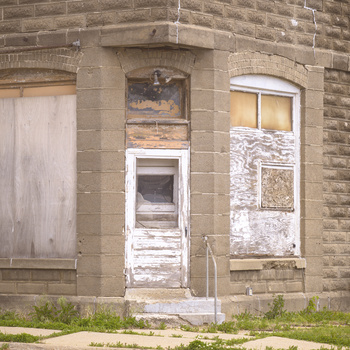 Weathered brick facade with boarded white door and window, showing decay and abandonment.