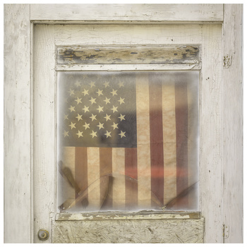Faded American flag with stars and stripes visible through weathered wooden frame window.