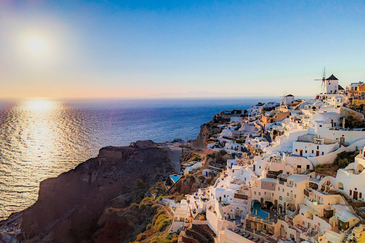 Whitewashed buildings cascade down a clifftop toward a golden-hour sea, with a windmill visible on the right.