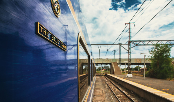 Blue passenger train car at a platform with railroad tracks and overhead electrical lines visible.