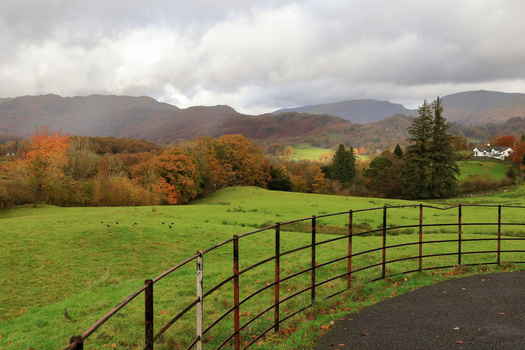 Black metal farm gate with rolling green pasture, autumn trees, and mountains under cloudy sky.