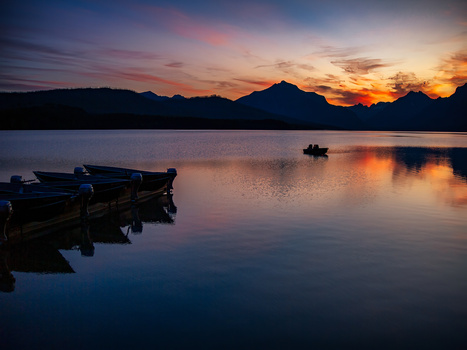 Fishing boats silhouetted against a vibrant sunset reflected in calm water, with mountains in the distance.