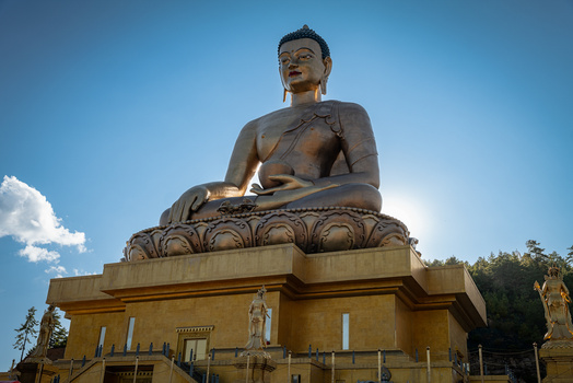 Large bronze Buddha statue seated on an ornate lotus-pedestal base, photographed against a clear blue sky.