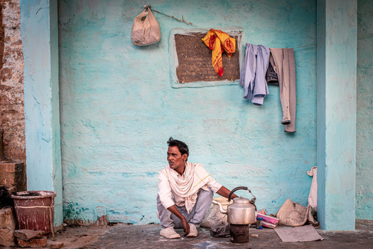 Man crouching beside cooking vessels against a weathered turquoise wall with hanging textiles.