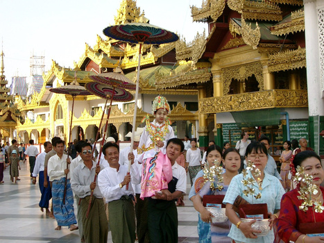 Procession of devotees in white and traditional dress walking past ornate golden temple structures with tiered roofs.