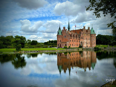 Brick castle with green spires reflected in still water, surrounded by green fields and trees under partly cloudy sky.