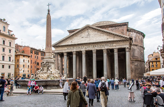 Historic Roman temple with Corinthian columns and obelisk in plaza crowded with tourists.