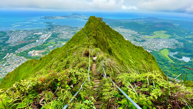 Steep hiking trail ascending a verdant volcanic peak with aerial view of coastal town and ocean below.