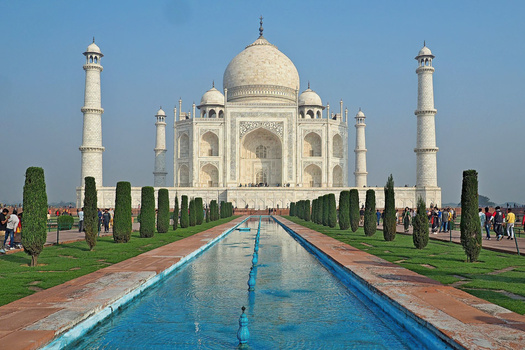 Symmetrical view of a marble mausoleum with a central dome and four minarets, flanked by cypress trees and a reflecting pool.