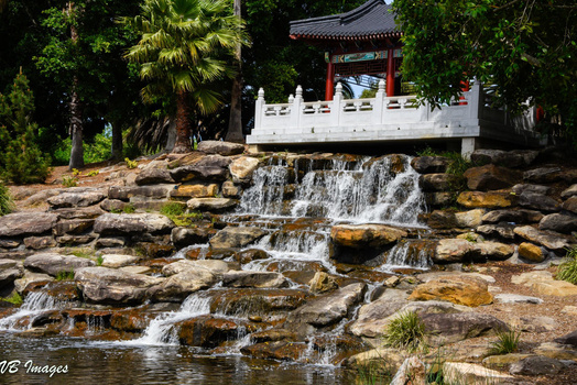 Cascading waterfall flowing over layered rocks with a traditional Asian pavilion in the background surrounded by lush trees.