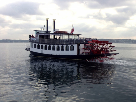 Classic paddle-wheel steamboat with white upper decks and red stern wheel on calm water under cloudy sky.