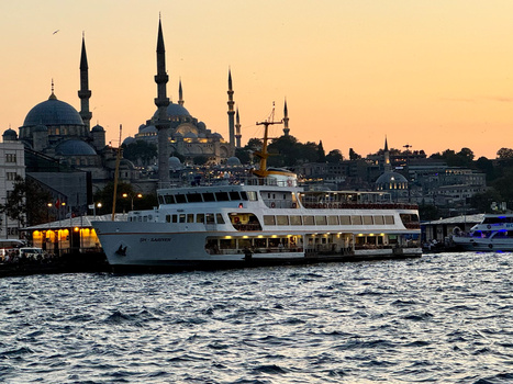 White passenger ferry on water at sunset with mosques and minarets silhouetted against golden sky.