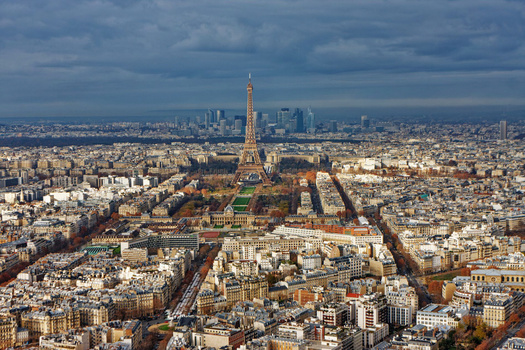 Aerial view of Paris with the Eiffel Tower dominating the center, surrounded by dense urban buildings and boulevards extending to the distant skyline.