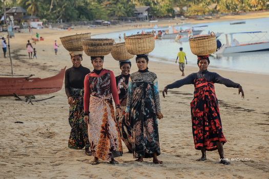 Five women in colorful patterned clothing stand on a beach carrying woven baskets on their heads.