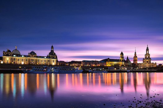 Illuminated historic buildings reflected in still water during twilight with purple and pink sky.