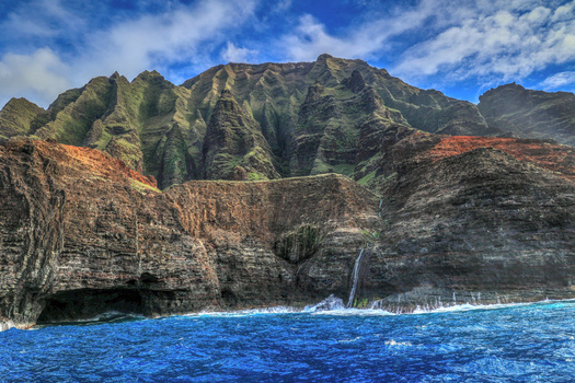 Steep sea cliffs with vertical green ridges rise from bright blue ocean water, with a small waterfall visible on the rocky face.