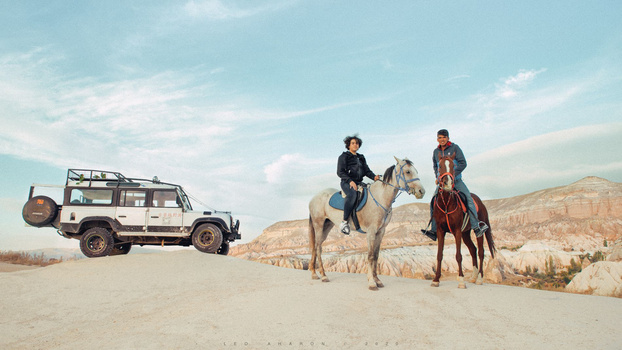 Two people ride horses in desert landscape next to a loaded Land Rover vehicle.